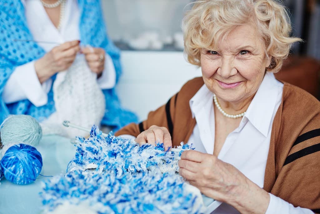 Portret de femeie senior cu părul blond, care se uită la cameră cu ochi negri adânci, în timp ce ține în mână o eșarfă tricotată neterminată, una dintre activități pentru vârstnici.