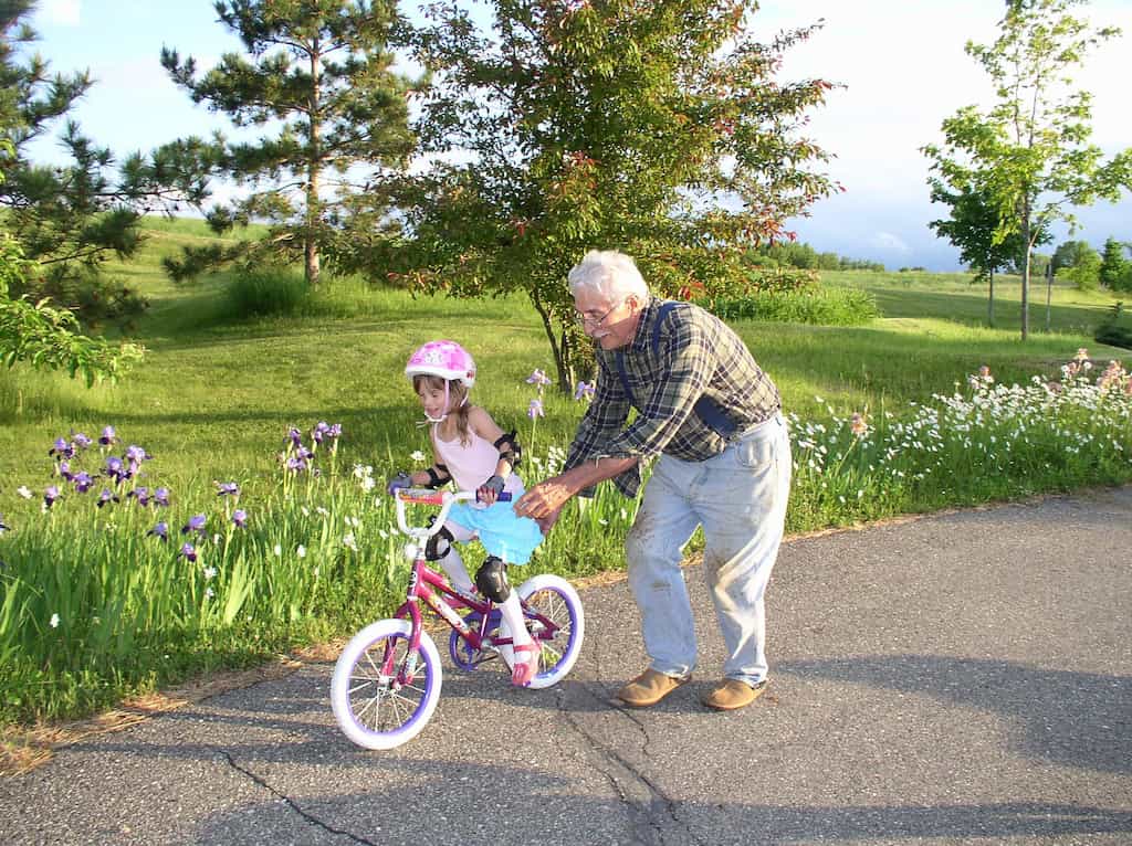 Bunicul învață pe nepoții să dea cu bicicleta, vara, fetiță pe o bicicletă cu bunicul care o ajută.