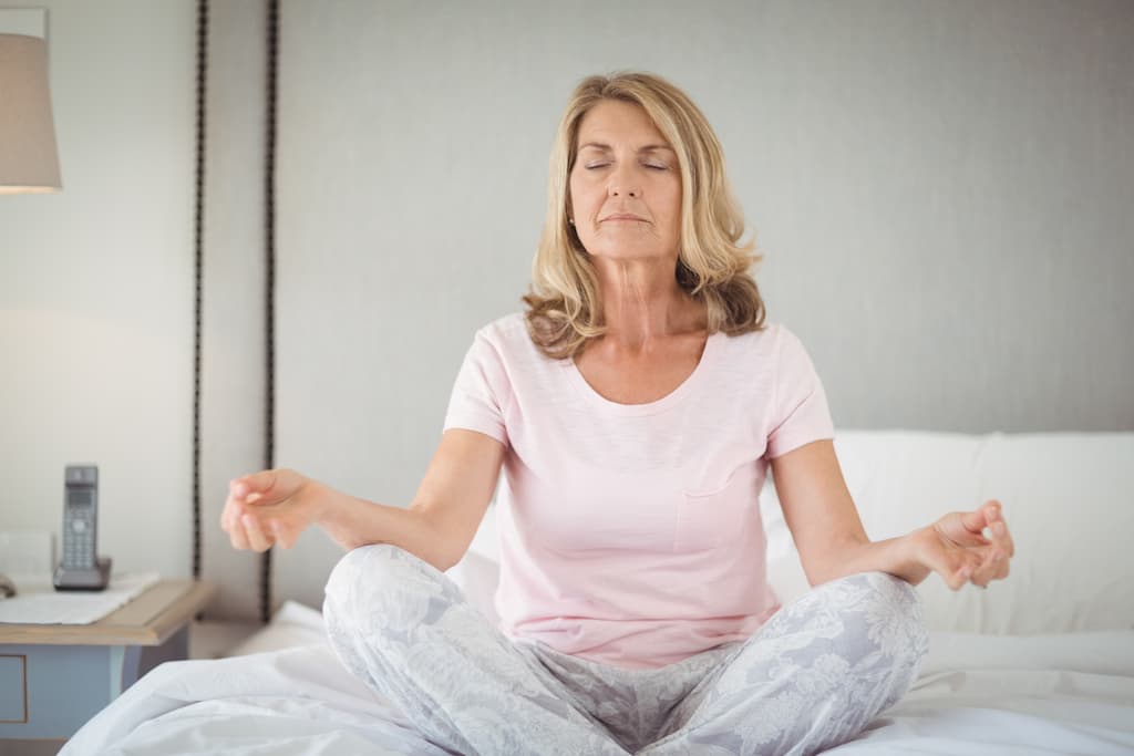 O doamnă în vârstă practică respirația de yoga pe pat cu ochii închiși în poziție lotus.