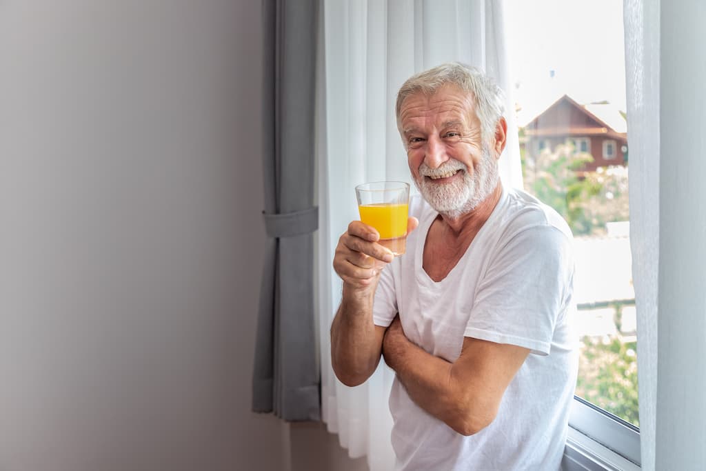 Sănătatea bărbaților peste 50 ani, bărbat senior cu barbă albă în tricou alb ține în mână un pahar cu suc de portocale zâmbind stă în picioare în fața geamului.